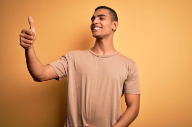 Sarı arka planda dikilen, gururlu, gülümseyen ve baş parmak hareketi yapan sıradan tişörtlü Afro-Amerikan genç adam.
