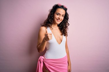 Tatilde kıvırcık saçlı güzel bir kadın pembe arka planda beyaz mayo giyerek mutlu bir el hareketi yapıyor. Kameraya bakıp başarıyı gösteren ifade onaylanıyor.