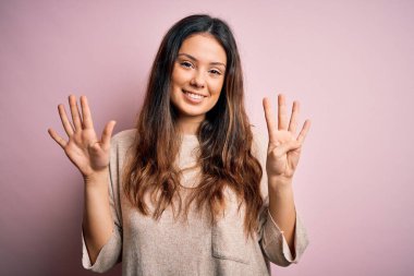Genç, güzel esmer kadın pembe arka planda dikilip dokuz numarayı gösterirken kendine güvenen ve mutlu gülümseyen..