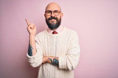 Sakallı, dövmeli, gözlüklü, pembe arka planda süveter giyen, yüzünde kocaman bir gülümseme olan, eli ve parmağı kameraya bakan yakışıklı kel bir adam..