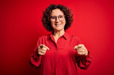 Orta yaşlı, kıvırcık saçlı, rahat gömlekli ve kırmızı arka planda gözlüklü güzel bir kadın mutlu ve komik yüzlü parmaklarını kameraya doğrultuyor. İyi enerji ve titreşimler.