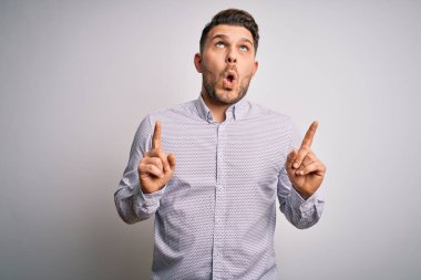 Mavi gözlü genç iş adamı şaşırmış ve şaşırmış bir şekilde yukarı bakıyor ve parmaklarını gösterip kollarını kaldırıyor..