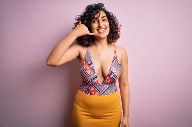 Tatilde, mayo ve pembe arka planda güneş gözlüğü takmış, gülümseyen, el ve parmaklarıyla telefonla konuşan genç, güzel bir Arap kadın. İletişim kavramları.