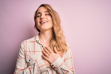 Genç, güzel, sarışın bir kadın pembe arka plan üzerinde dikiliyor elleri göğsünde, gözleri kapalı ve yüzünde minnet dolu bir jest ile gülümsüyor. Sağlık kavramı.