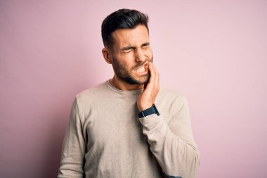İzole edilmiş pembe arka plan üzerinde dikilen genç yakışıklı bir adam diş ağrısı ya da diş hastalığından dolayı acı dolu bir ifadeyle ağıza dokunuyor. Dişçi