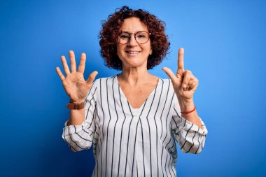 Orta yaşlı, kıvırcık saçlı, rahat çizgili tişört giyen güzel bir kadın izole arka planda 7 numaralı parmaklarını gösterip gülümserken mutlu ve kendinden emin..