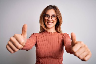 Genç ve güzel esmer kadın, günlük süveter ve gözlük takıp beyaz arka planda olumlu jest yapmayı onaylıyor. Elleriyle gülümsüyor ve başarı için seviniyor. Kazanan jest.