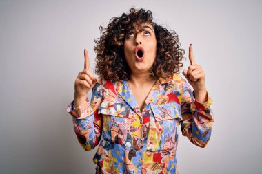 Genç ve güzel kıvırcık Arap kadın çiçek rengi gömlek giyiyor beyaz arka planda duruyor şaşkın şaşkın yukarı bakıyor ve parmaklarıyla işaret ediyor ve kollarını kaldırıyor..