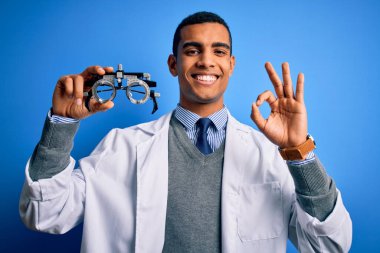 Yakışıklı Afro-Amerikan optik adam mavi arka planda optometri gözlüğü tutuyor. Parmaklarıyla imza atıyor. Mükemmel bir sembol.
