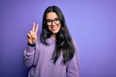 Genç esmer kadın mor renkli gözlük takıyor. İzole edilmiş arka planda. İki numaralı parmaklarıyla işaret ediyor ve gülümsüyor..