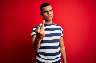 Handsome african american man wearing casual striped t-shirt standing over red background Showing middle finger, impolite and rude fuck off expression