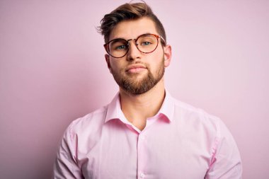 Sakallı, mavi gözlü, pembe gömlekli ve gözlüklü yakışıklı sarışın adam. Yüzünde ciddi bir ifadeyle rahatlamış. Basit ve doğal bir şekilde kameraya bakıyorsun..