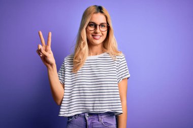 Genç, sarışın, çizgili tişört ve gözlük takan mor arka planda iki numaralı parmaklarıyla kendini beğenmiş ve mutlu gülümseyen bir kadın..