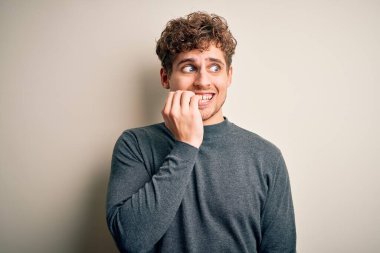 Sarışın, kıvırcık saçlı, beyaz arka planda sıradan süveter giyen, elleri ağız yiyen, gergin ve gergin genç bir adam. Endişe sorunu.