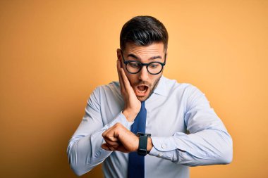 Sarı arka planda duran kravat ve gözlük takan genç yakışıklı işadamı, saati endişeli, geç kalmaktan korkan saate bakıyor.