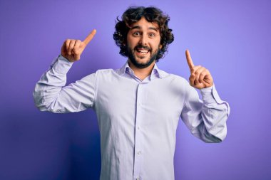 Mor arka plan üzerinde duran sakallı yakışıklı iş adamı. Şaşırmış, şaşırmış ve parmaklarıyla işaret ediyor ve kollarını kaldırıyordu..
