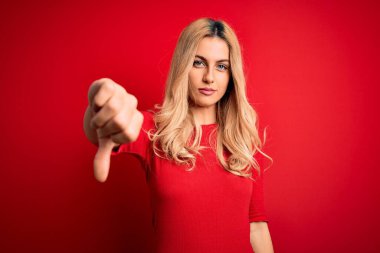 İzole edilmiş kırmızı arka plan üzerinde duran gündelik tişört giyen genç, güzel sarışın kadın mutsuz, reddedilmiş ve olumsuz hareketler sergiliyor. Kötü ifade.