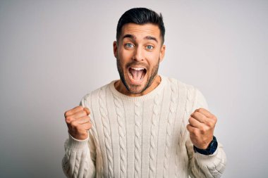 İzole edilmiş beyaz arka plan üzerinde duran sıradan süveter giyen genç yakışıklı adam kollarını kaldırarak ve açık gözlerle başarının şaşkınlığını ve şaşkınlığını kutluyor. Kazanan konsept.
