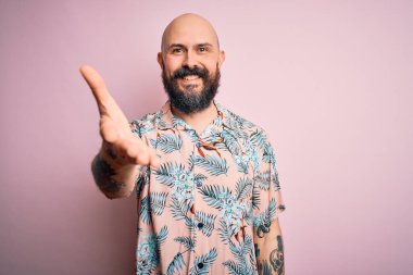 Sakallı ve dövmeli yakışıklı, pembe arka planda sıradan bir çiçek gömleği giyen, dostça selamlaşmak ve el sıkışmak için gülümseyen kel bir adam. Başarılı bir iş..