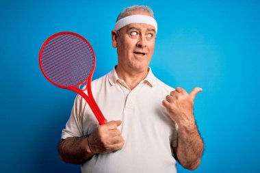 Tenis oynayan orta yaşlı, ağarmış bir sporcu izole edilmiş mavi arka planda raket kullanıyor ve gülümseyen bir yüzle baş parmağıyla işaret ediyor.