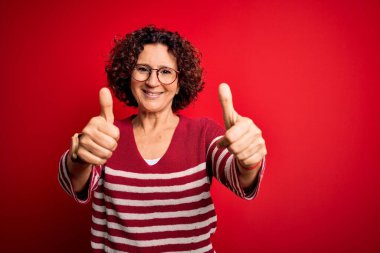 Orta yaşlı güzel kıvırcık saçlı kadın kırmızı arka planda sıradan çizgili süveter giyiyor. Elle olumlu jest yapmayı onaylıyor. Gülümsüyor ve başarı için seviniyor. Kazanan jest.