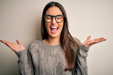 Genç, güzel esmer kadın, beyaz arka plan üzerine gündelik süveter ve gözlük takmış, çılgın ve çılgın başarısını kutluyor, kolları havada ve gözleri kapalı, heyecanlı bir şekilde çığlık atıyor. Kazanan konsept