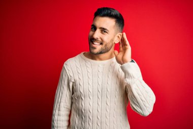 İzole edilmiş kırmızı arka planda dikilen, gündelik beyaz süveter giyen genç yakışıklı bir adam. Kulağı kulağında, dedikoduları ve dedikoduları dinliyor. Sağırlık kavramı.