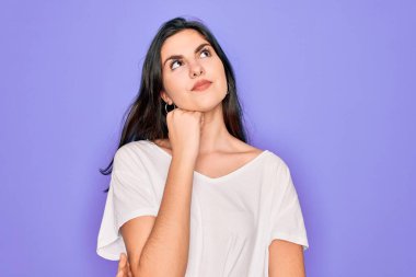 Genç, güzel, esmer bir kadın, mor arka plan üzerine gündelik beyaz tişört giymiş, elleri çenesinde soru soran, düşünceli bir ifade takınıyor. Gülen ve düşünceli bir yüz. Şüphe kavramı.