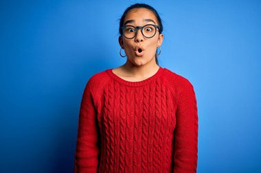 Genç ve güzel Asyalı kadın mavi arka planda günlük süveter ve gözlük takıyor. Şaşırmış ve korkmuş bir ifade, korku ve heyecanlı bir yüz ifadesi var..