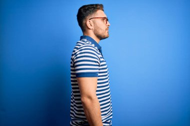 Mavi gözlü, gözlüklü ve mavi arkaplanlı sıradan çizgili tişörtlü, rahat bir görünüm takınan, kendine güvenen bir gülümsemesi olan genç bir adam..