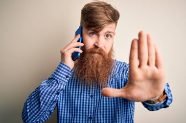 Kızıl saçlı, sakallı İrlandalı bir adam, kapalı bir arka plan üzerinden akıllı telefondan konuşuyor. Eli açık, ciddi ve kendinden emin bir ifadeyle dur işareti yapıyor, savunma jesti yapıyor.