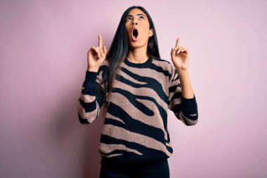 Pembe arka planda hayvan desenli süveter giyen genç ve güzel İspanyol kadın hayret ve şaşkınlık içinde yukarı baktı ve parmaklarıyla işaret etti ve kolunu kaldırdı..