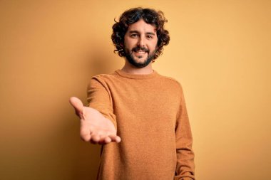 Sakallı, sıradan bir süveter giyen, sarı arka planda durup gülümseyen, yardım eli uzatan ve kabul gören genç yakışıklı bir adam..