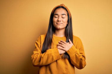 Sarı arka plan üzerine gündelik süveter ve diyabet giyen genç, güzel Asyalı kadın elleri göğsünde, gözleri kapalı ve yüzünde minnet dolu bir jest ile gülümsüyor. Sağlık kavramı.