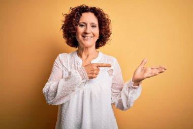 Orta yaşlı güzel kıvırcık saçlı kadın sarı arka planda rahat bir yaz elbisesi giyiyor kameraya hayret ediyor ve gülümsüyor..