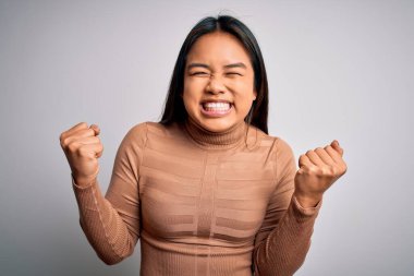 Genç ve güzel Asyalı kız balıkçı yaka kazak giyerek beyaz arka planda mutlu ve heyecanlı bir şekilde kolları havada, gülümseyerek ve başarı için çığlık atarak zafer hareketi yapıyor. Kutlama konsepti.