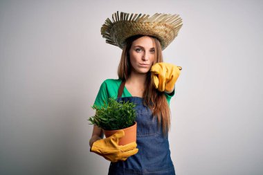 Önlük ve şapka takan genç, kızıl saçlı bir kadın elinde bir saksı, kamerayı işaret ediyor ve sen, el işareti, ön taraftan pozitif ve kendinden emin bir jest.