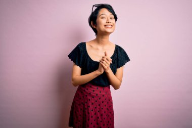 Genç ve güzel Asyalı bir kız pembe arka plan üzerinde rahat ve neşeli bir şekilde gülümseyerek ellerini çapraz yaparak dikiliyor. Başarı ve iyimser