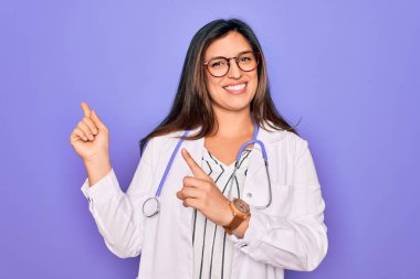 Mor arka planda steteskop ve tıbbi ceket giyen profesyonel doktor kadın gülümseyip kameraya bakıyor ve iki eliyle ve parmaklarıyla yan tarafa bakıyor..