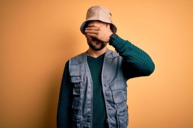 Tatilde sakallı yakışıklı turist sarı arka planda kaşif şapkası takıyor gözlerini eliyle kapatıyor, ciddi ve üzgün görünüyor. Görme, saklama ve reddetme konsepti