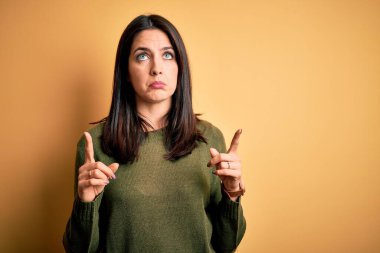 Sarı arka planda yeşil süveter giyen, mavi gözlü genç esmer kadın üzgün ve üzgün görünüyor, parmaklarıyla yön gösteriyor, mutsuz ve depresif..