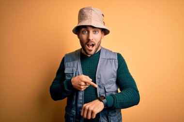 Tatilde sakallı yakışıklı bir turist sarı arka planda kaşif şapkası takıyor. Aceleyle saati, sabırsızlığı ve gecikmeyi gösteriyor.