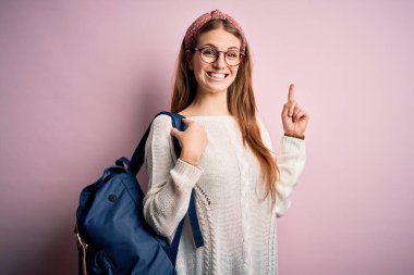 Sırt çantası ve pembe arka planda gözlük takan güzel kızıl saçlı bayan bir fikir ya da soru işareti ile şaşırdı.