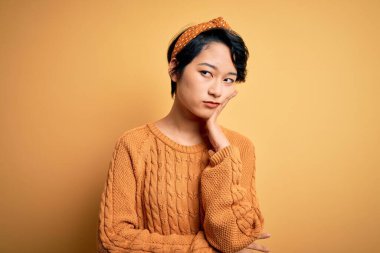 Genç, güzel Asyalı bir kız gündelik süveter ve Diadem giyip sarı arka planda duruyor. Kollarını kavuşturup depresyondan sıkılmış ve yorgun görünüyor..