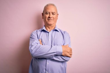 Orta yaşlı yakışıklı, beyaz gömlekli, pembe arka planda duran mutlu yüzlü, kollarını kavuşturup kameraya bakan bir adam. Pozitif kişi.