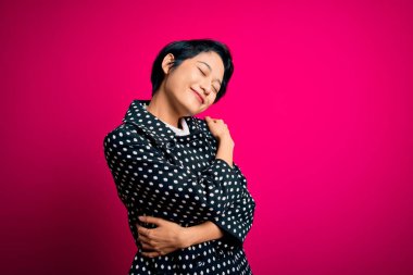 Genç, güzel Asyalı bir kız, günlük ceket giyiyor pembe arka planda dikiliyor, kendine sarılıyor, mutlu ve pozitif, kendinden emin gülümsüyor. Sevgi ve özgüven.