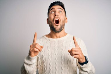 İzole edilmiş beyaz arka plan üzerinde duran gündelik süveter giyen genç yakışıklı adam şaşırmış ve şaşırmış bir şekilde yukarı bakıyor ve parmaklarıyla işaret ediyor ve kolunu kaldırıyor..
