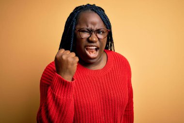 Afro-Amerikalı, örgülü, sarı arka plan üzerine normal süveter giyen kızgın, kızgın, sinirli ve öfkeliyken öfkeyle bağıran bir kadın. Öfke ve agresif kavram.