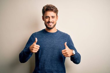 Sakallı, sıradan bir süveter giyen, beyaz arka plan başarı tabelasının üzerinde duran eli ile olumlu jest yapan, gülümseyen ve mutlu genç bir adam. Neşeli ifade ve kazanan jest.