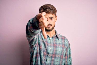 Sakallı, pembe arka planda sıradan bir gömlek giyen, mutsuz, sinirli, reddedilmiş ve olumsuz jestli genç bir adam. Kötü ifade.